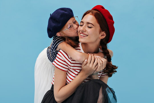 Ginger cute girl with freckles in dark beret and moern lothes kisses in cheek and hugs her elder smiling sister in red hat..