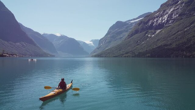 Woman kayaking turquoise Lovatnet lake during sunny summer day - Backward moving aerial close to water with kayak moving towards camera - Beautiful Norway mountain landscape with glacier in background