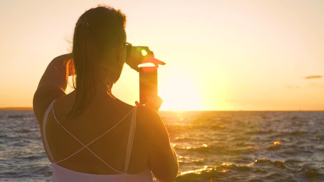 SLOW MOTION, CLOSE UP, LENS FLARE, DOF: Female traveler takes photos of the sun setting behind Adriatic sea. Young woman on relaxing summer vacation in Zadar is taking photos of the golden sunset.