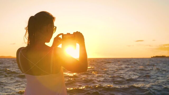 SLOW MOTION, CLOSE UP, LENS FLARE, DOF: Young woman on relaxing summer vacation in Zadar is taking photos of the golden sunset. Female traveler takes photos of the sun setting behind Adriatic sea.