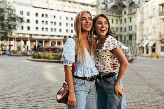 Blonde and brunette women in stylish outfits look away in good mood. Charming girls in floral tops and jeans smile, hug and pose outside.