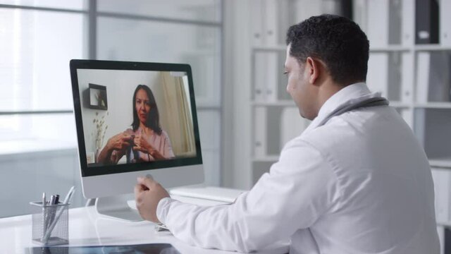Doctor sitting at desk in front of computer monitor watching his female patient checking blood pressure