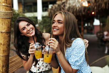 Cool young blonde lady in blue top and tanned brunette curly girl in white blouse smile widely, walk outside and drink tasty lemonade.