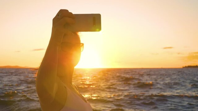 SLOW MOTION, CLOSE UP, LENS FLARE, DOF: Smiling young Caucasian female tourist on relaxing summer vacation in Zadar, Croatia takes selfies in front of the calm Adriatic sea at idyllic golden sunset.