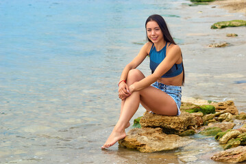 Fototapeta na wymiar Young slender girl at the seaside resort. Woman in denim shorts sitting by the sea.