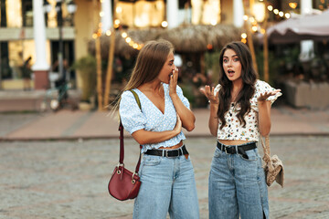 Curly brunette woman in white floral top and jeans talks to her friend and tells her secrets. Blonde tanned girl in denim pants and blue blouse looks surprised and covers mouth. Women walks in city.