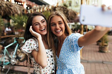 Cheerful girlfriends in great mood take selfie outdoors and smile. Pretty blonde woman in trendy blue blouse holds cellphone.