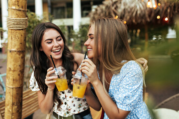 Good-humored optimistic brunette woman in white floral top and happy blonde tanned girl in blue blouse talk, smile and drink lemonade outside.