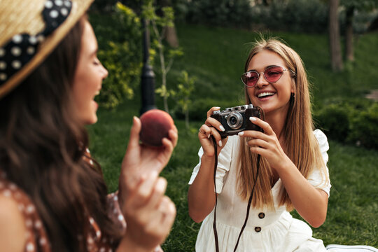 Cheerful girlfriends in stylish summer dresses smile and have picnic outside. Cool blonde lady in red sunglasses takes photo of brunette woman holding apple.