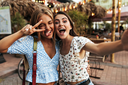 Tanned blonde girl in blue blouse demonstrates tongue, shows peace sign and winked. Beautiful brunette woman in floral top takes selfie with attractive friend outside.