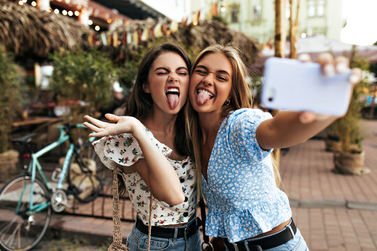 Active young girls in stylish blouses make funny faces, show tongues and take selfie. Pretty blonde woman in blue top holds purple phone.