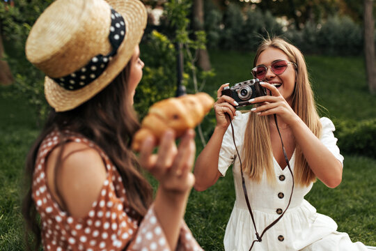 Tanned blonde woman in white dress and red sunglasses takes photo of her brunette curly friend using retro camera. Lady in polka dot dress and boater holds croissant.