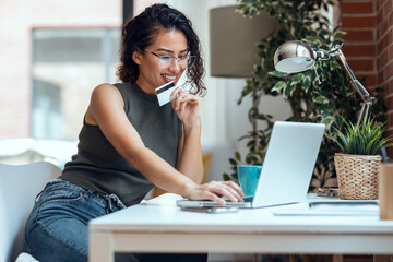 Cute woman holding white credit card for shopping online with computer while sitting in living room...