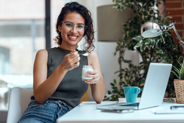 Beautiful young business woman working with laptop while eating yogurt looking at camera in living...