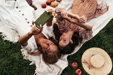 Pretty brunette and blonde women in summer dresses lay on linen rug and take selfie. Girl in white outfit holds phone. Ladies have picnic on grass.