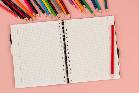 Open school squared notebook and color pencils on the pink background. Blank white sheet of paper book on the table. Office supplies on the desktop. Back to school. Copy space for text. Top view