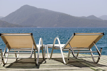 Sea resort, two empty deck chairs on a wooden pier on green mountains background. Beach vacation on picturesque nature