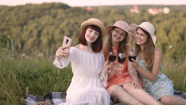 People and friendship concept. Cheerful company of three female friends, having fun and enjoying summer picnic and toasting glasses with wine, making selfie photo on smartphone