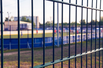 Through the bars you can see the sports stadium where the guys train. Focus is on the grille, and the rear soccer field is blurry. The footballers of the young team are playing football.