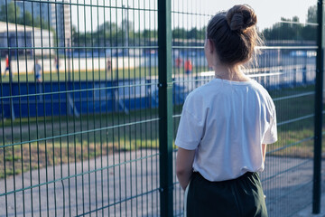 A girl stands and looks through the fence at the athletes playing at the stadium. The girl wants, but cannot play sports.