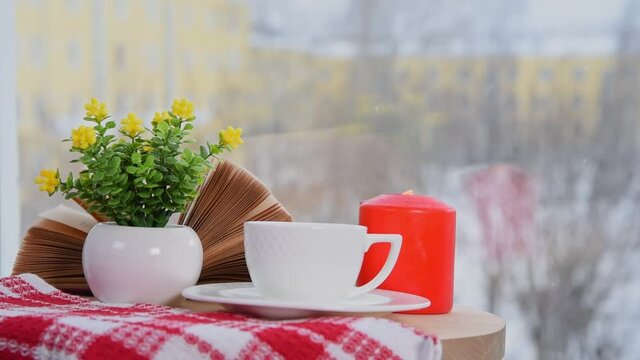 White coffee cup, book, burning candle and vase with flowers on table in cafe. Winter time, It snows outside. Coziness atmosphere