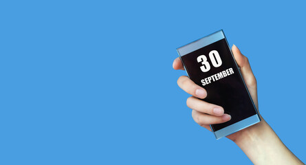 september 30. 30th day of the month, calendar date.Woman's hand holds mobile phone with blank screen on blue isolated background.Autumn month, day of the year concept