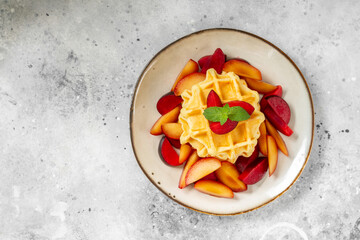 Traditional classic Belgian or Brussels waffles with red plums in a ceramic plate on a light gray kitchen table top view	