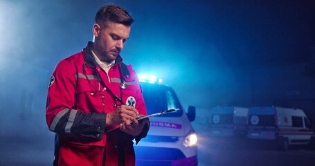 Handsome young Caucasian male paramedic in red uniform writing with pen in hand in folder with...
