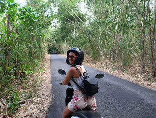 Young happy Caucasian woman riding a rental motorcycle with safety helmet and sunglasses to drive safely on the trip around the island of Bali visiting a beautiful bamboo forest