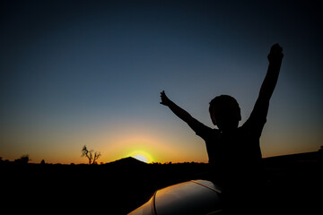 Silhouette of child with arms up on top of a vehicle in front of Pyramid Hill in Victoria, Australia at sunset