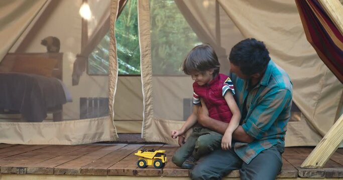 Father plays with his son a toy dump truck in a large tent in nature. They sit on a wooden floor, and the walls of the house develop around them. Outside the window is the forest. Man holding son