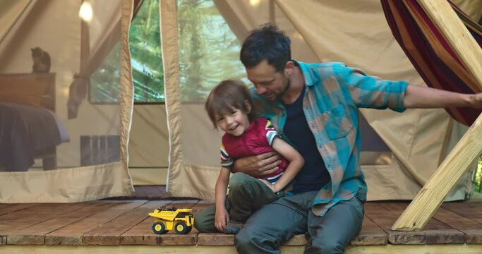 A handsome man with a beard, wearing a shirt, plays with his handsome son. They sit in a large tent in nature and have fun. A toy dump truck stands next to them. Outside the window is the forest