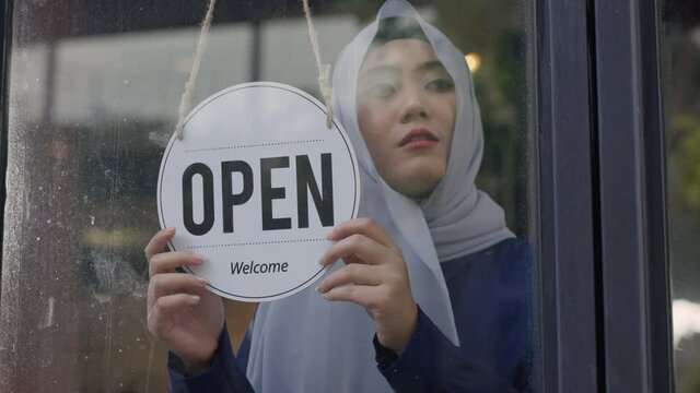 Young Asian Muslim woman wearing hijab, turning sign to closed the coffee shop or restaurant due to coronavirus crisis. Female Muslim business owner changing sign from open to closed.