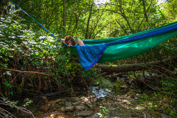 A girl lies resting in a green hammock stretched out on blue slings over a small mountain river in...