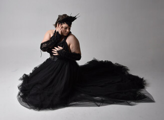 Full length portrait of young  plus sized woman with short  hair,  wearing long black tuille gothic gown with crown and gloves, sitting pose on light studio background.