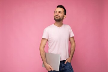 Handsome happy young unshaven man holding laptop computer looking up in t-shirt on isolated pink background