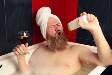 Cheerful bearded man taking bubble bath holding glass of red wine and singing into shampoo bottle with facial expression. Relaxation at home after hard day. Happy guy enjoying life.