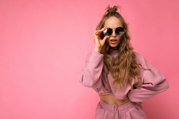 Shot of beautiful dissatisfied asking young dark blonde curly woman isolated over pink background wall wearing casual pink sport clothes and stylish sunglasses looking at camera and having questions
