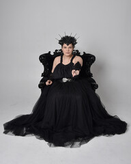 Full length portrait of young  plus sized woman with short  hair,  wearing long black tulle gothic gown with crown and gloves, sitting pose on a ornate throne chair on light studio background.