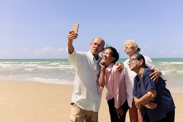 Group of four elderly old senior friends traveling outdoor together, take a selfie on mobile phone, have fun and enjoy spending time on sand beach, resting and relaxing on holiday vacation.