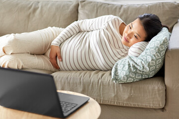 pregnancy, rest, people and expectation concept - happy smiling pregnant asian woman with laptop computer sitting on sofa at home