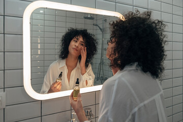 Young mixed race female gently applying face oil serum with fingers in modern bathroom.