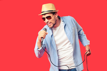 A white man in a hat and sunglasses sings a song into a microphone. Vocal performance of the song. Advertising background for a karaoke club or bar. An emotional singer. Studio portrait on a red
