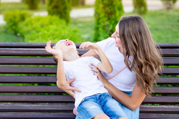 happy mom with her baby son in the park on a bench in the summer have fun hugging, laughing and playing