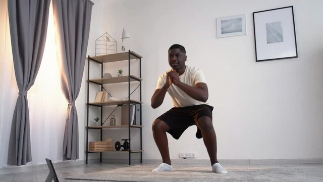 Home training. Happy black man. Enjoying sport. Healthy lifestyle. Smiling african guy casual cloth doing deep squats workout looking portable computer on floor light room interior.