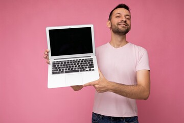 Handsome smiling brunet man holding laptop computer looking at camera in t-shirt on isolated pink background