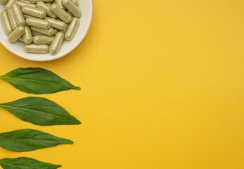 Fototapeta na wymiar Andrographis paniculata capsules are in a white dish and green leaves isolated on yellow background. Space for text. Herbal, medicine, and healthcare concept