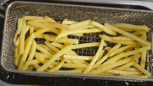 Top view of male cook shaking a lattice with french fries being prepared in hot boiling oil. Chef lowering a grid with potatoes into deep fryer. Process of making fast food at cuisine. Slow motion