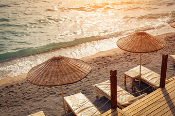 Empty sun bed chairs with mattress and straw beach umbrellas on beach during sunset in summer.