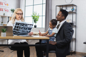 Handsome young african american father with his adorable little daughter listening to explanation of x-ray CT scan from confident female doctor pediatrician in modern medical office